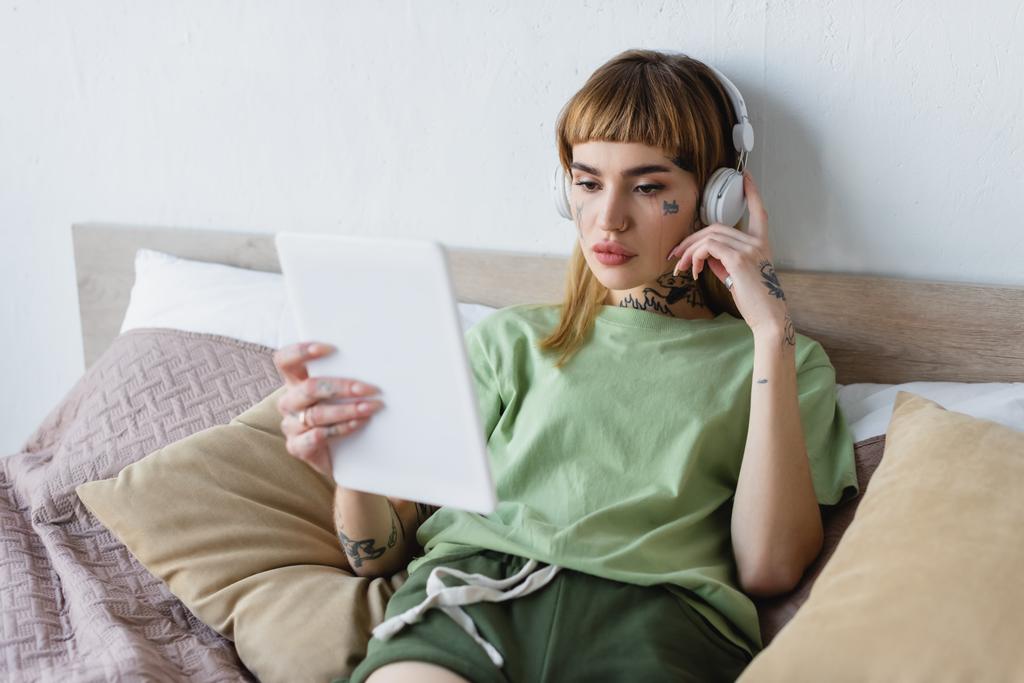 Kulaklıklı genç bir kadın yatak odasında podcast dinlerken bulanık dijital tablete bakıyor. - Fotoğraf, Görsel
