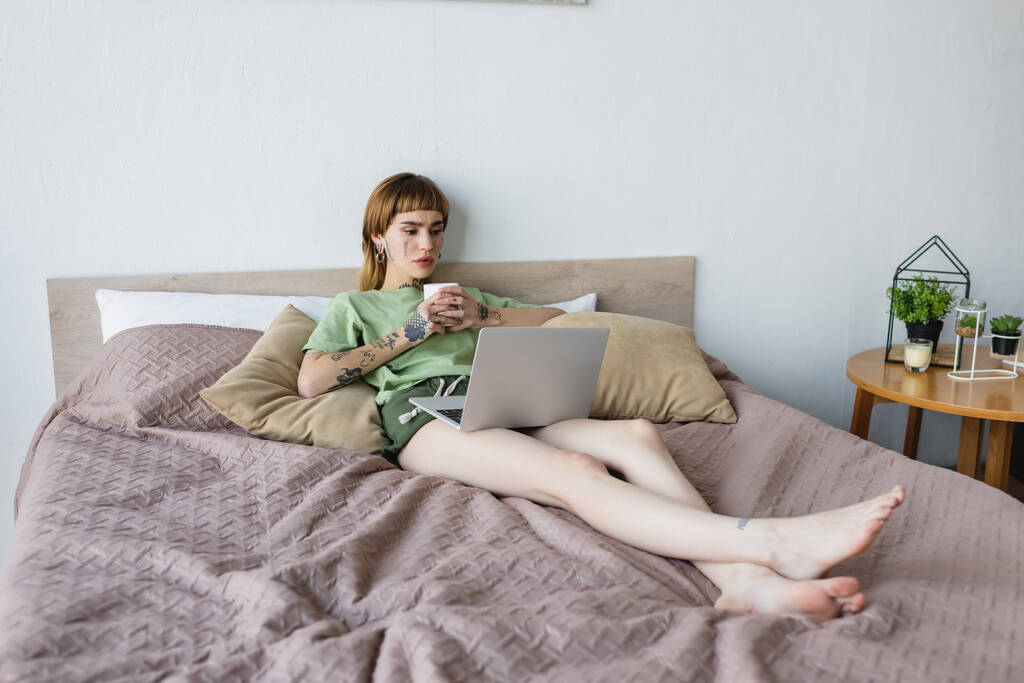 Ganzkörperansicht einer jungen tätowierten Frau, die Tee trinkt und sich Film auf Laptop im Bett ansieht - Foto, Bild
