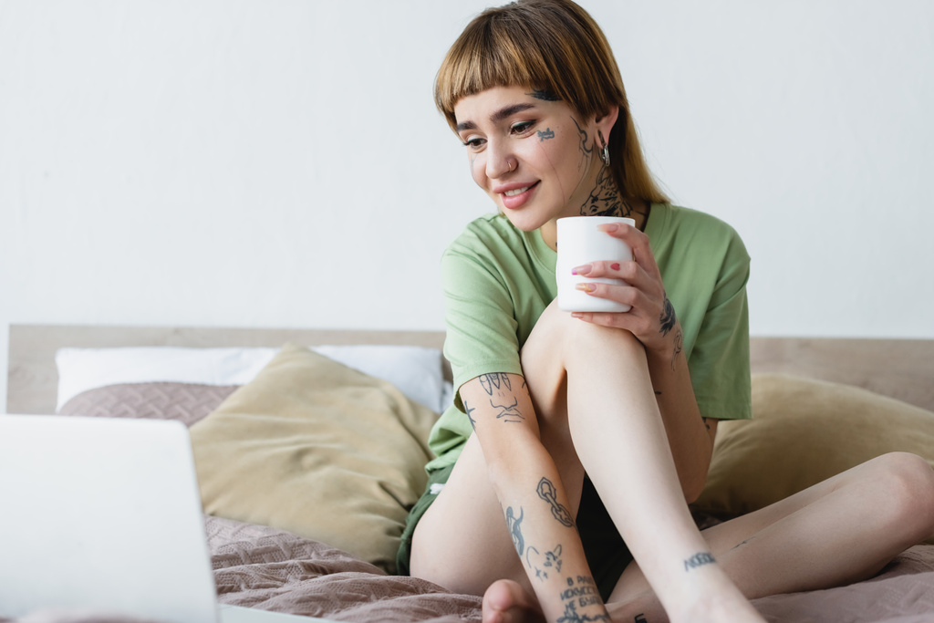 szczęśliwa kobieta z wytatuowanym ciałem trzymając filiżankę herbaty podczas oglądania filmu na niewyraźnym laptopie - Zdjęcie, obraz