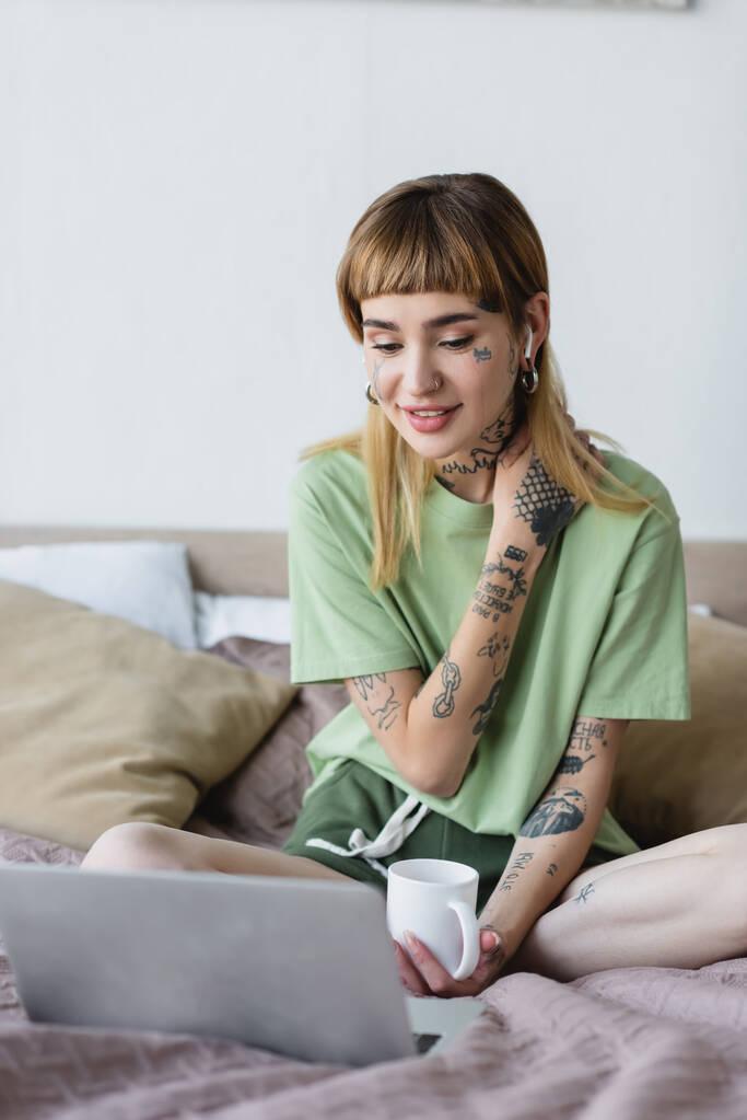 positive woman with tattooed body watching film on laptop in bedroom - Photo, Image