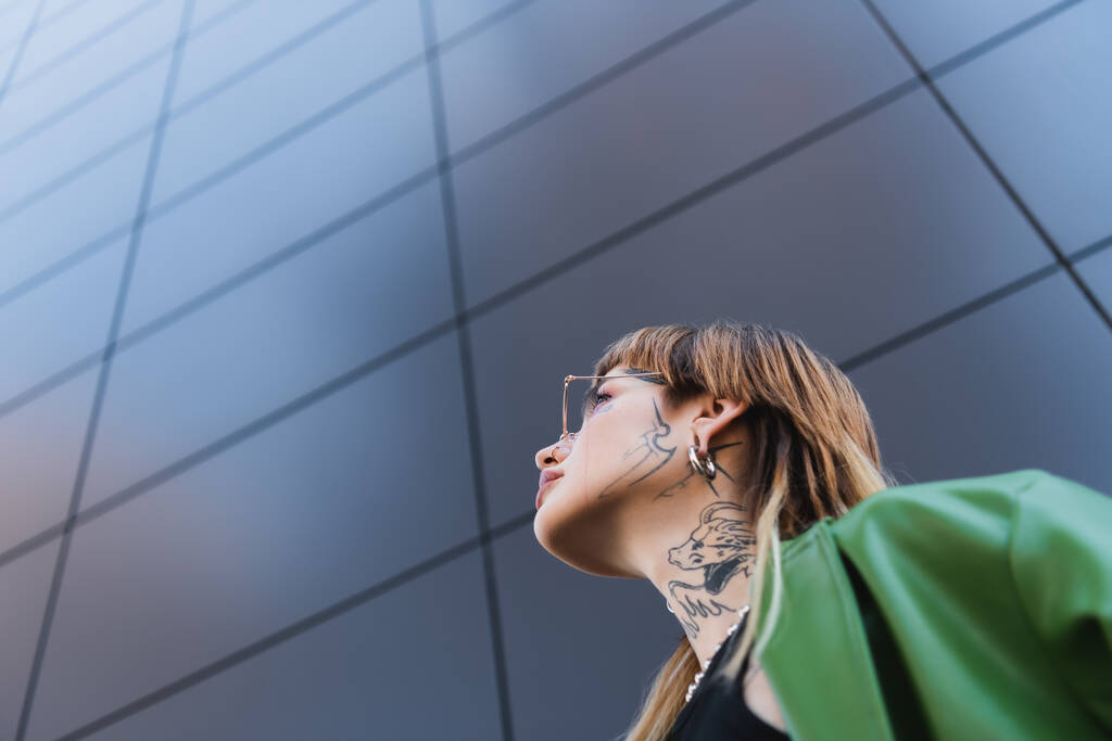 lage hoek uitzicht van stijlvolle getatoeëerde vrouw in de buurt van hoge grijze muur op stedelijke straat - Foto, afbeelding
