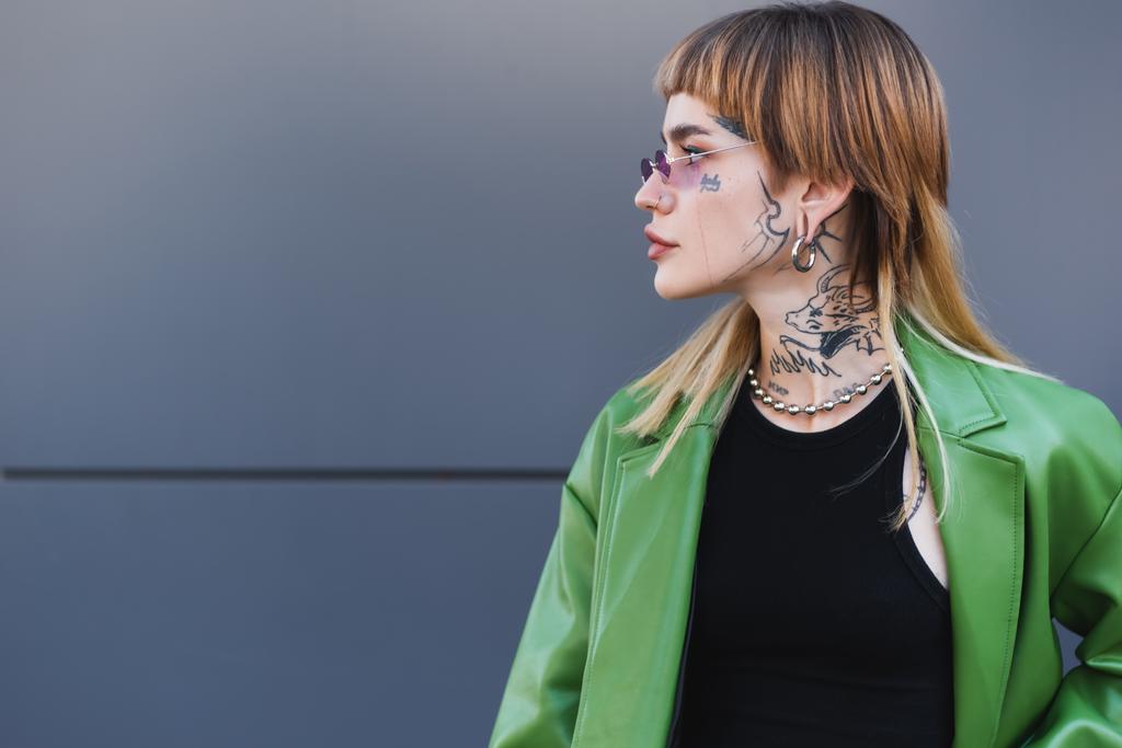 tattooed woman in stylish eyeglasses, green jacket and silver necklace looking away near grey wall - Photo, Image