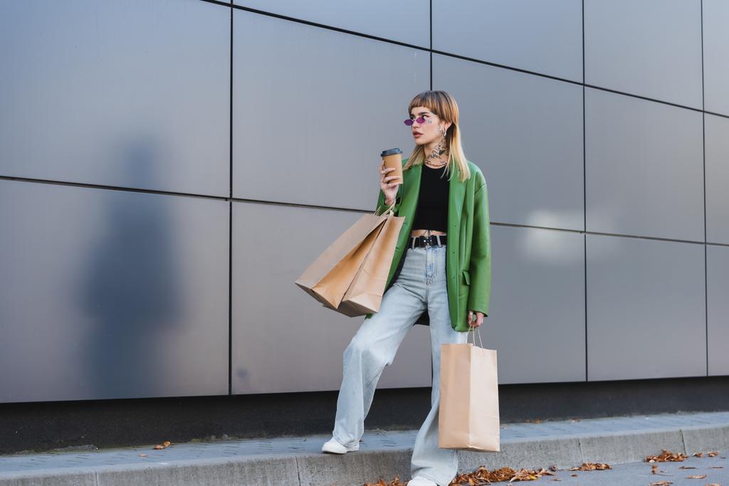 κομψή γυναίκα με τατουάζ σε πράσινο σακάκι και τζιν στέκεται με τσάντες για ψώνια και χάρτινο κύπελλο σε εξωτερικούς χώρους - Φωτογραφία, εικόνα