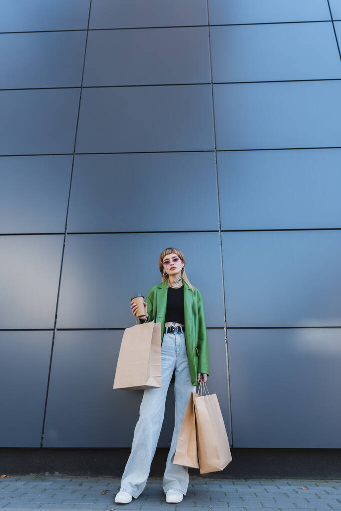 グレーの壁の近くに立っているジーンズのトレンディーな入れ墨の女性紙コップとショッピングバッグ - 写真・画像
