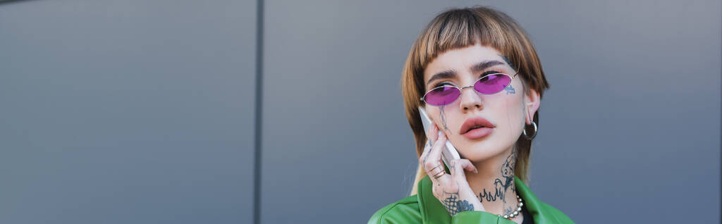 mulher tatuada em óculos elegantes falando no telefone celular perto da parede cinza, banner - Foto, Imagem