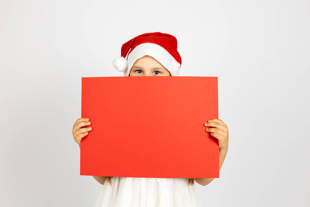 Noel Baba şapkalı mutlu bir kızın portresi ellerinde kırmızı boş bir posterin arkasına saklanıyor, beyaz arka planda izole edilmiş. - Fotoğraf, Görsel