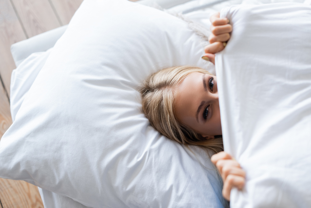 widok z góry młoda kobieta przykrywa twarz z kocem leżąc na łóżku - Zdjęcie, obraz