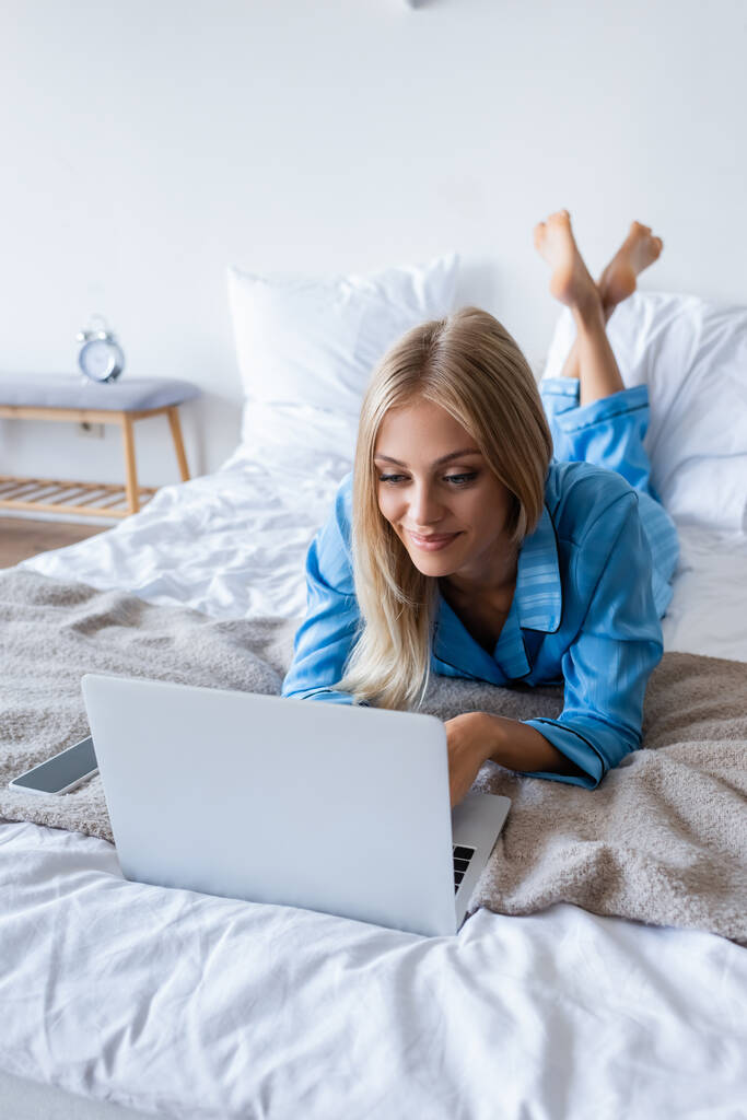 elégedett fiatal nő pizsama segítségével laptop a hálószobában  - Fotó, kép