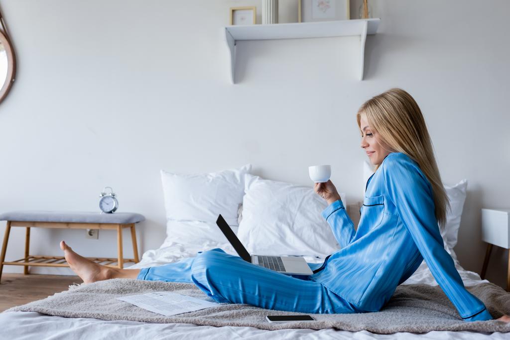 πλαϊνή άποψη του ευτυχισμένη νεαρή γυναίκα με πιτζάμες κοιτάζοντας φορητό υπολογιστή και κρατώντας φλιτζάνι καφέ  - Φωτογραφία, εικόνα