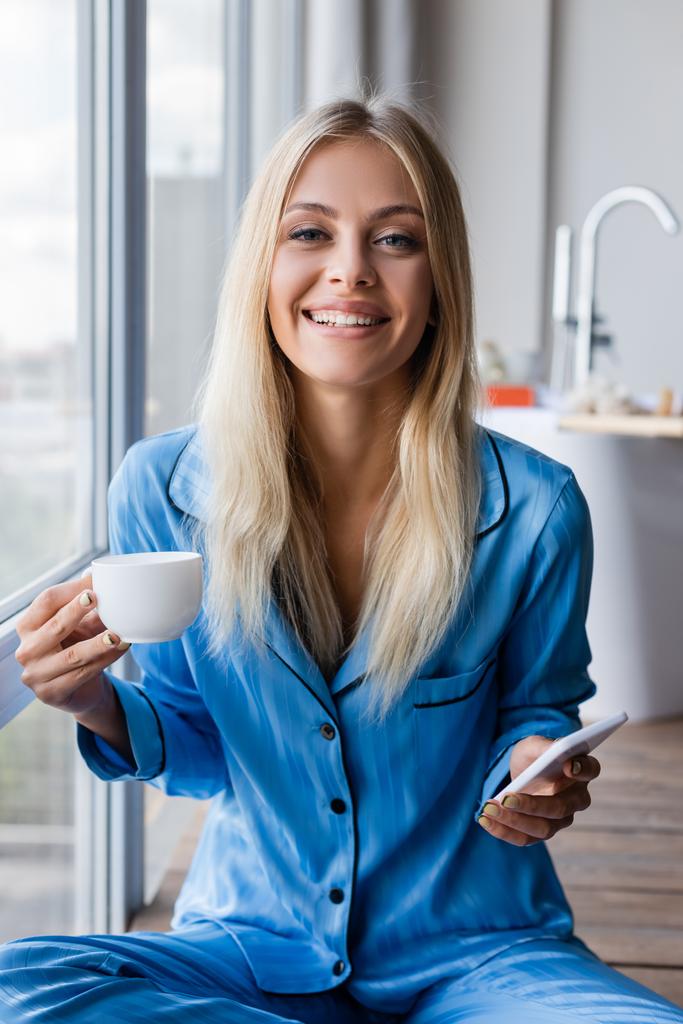Pencerenin yanında cep telefonu ve kahve tutan mutlu genç bir kadın.  - Fotoğraf, Görsel