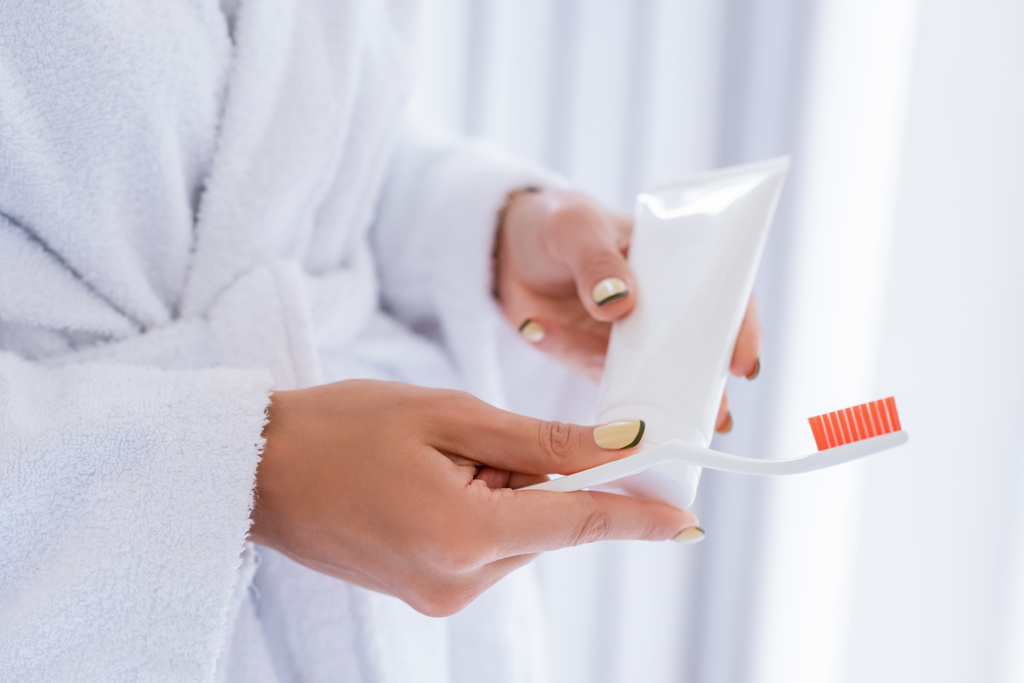 częściowy widok młodej kobiety trzymającej szczoteczkę do zębów i tubkę pasty do zębów w łazience - Zdjęcie, obraz