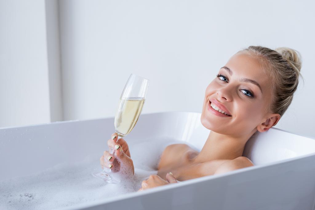 Genç ve neşeli bir kadın banyo yapıyor ve elinde bir kadeh şampanya tutuyor.  - Fotoğraf, Görsel