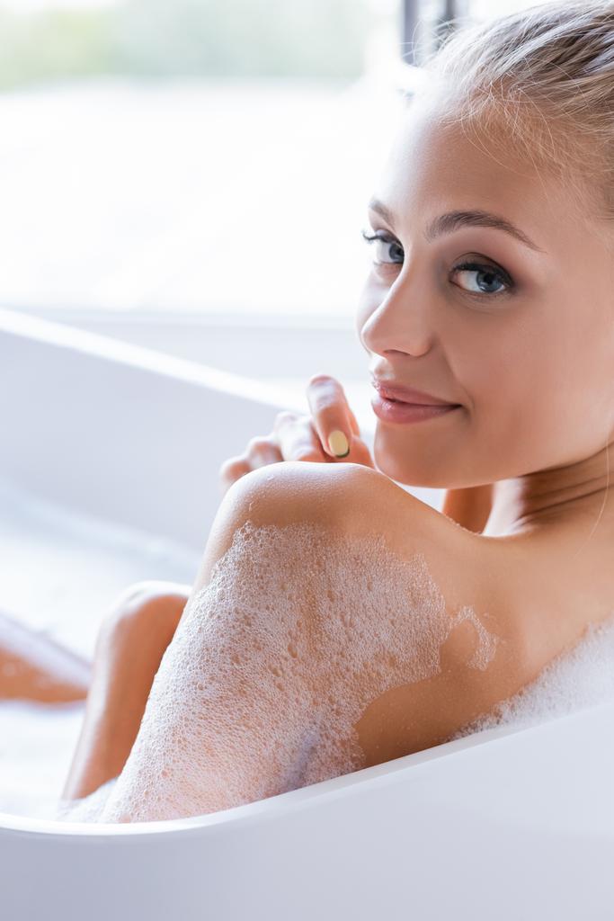 довольная молодая и мокрая женщина с пеной на теле принимающая ванну  - Фото, изображение