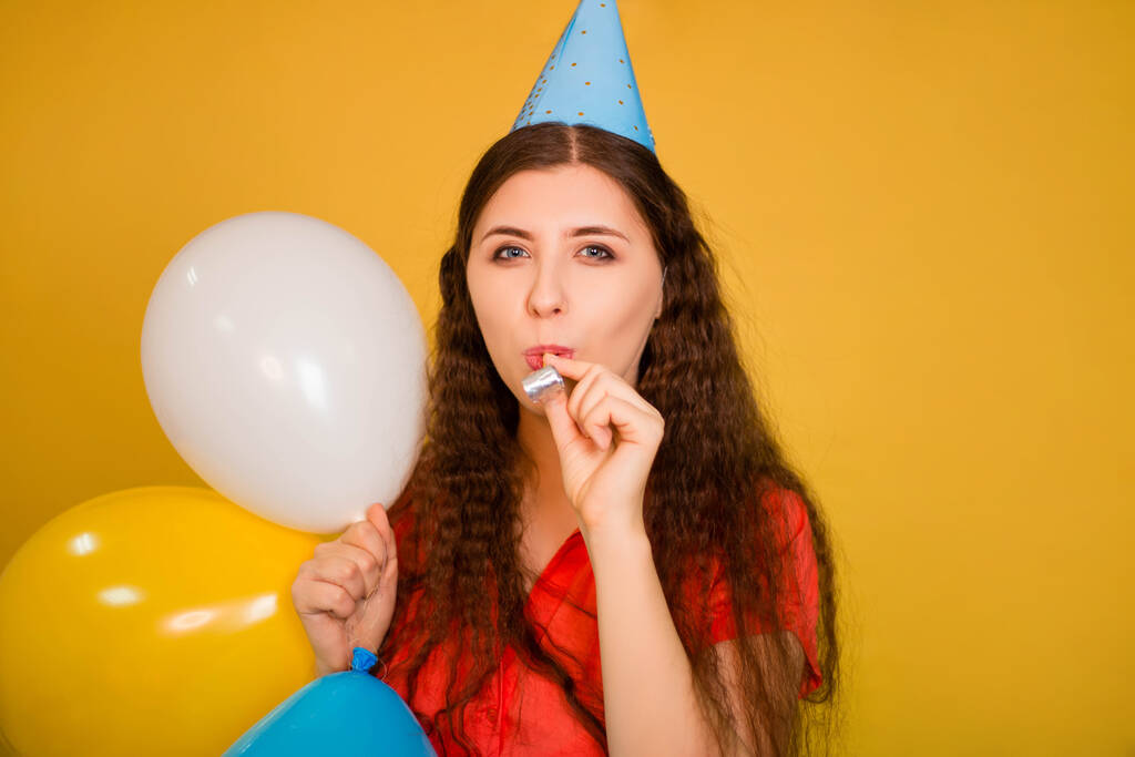 Portret van een jonge vrouw in een feestelijke pet die een pijp blaast met veelkleurige ballonnen in haar hand geïsoleerd op een gele achtergrond. lawaaierige verjaardag concept. - Foto, afbeelding