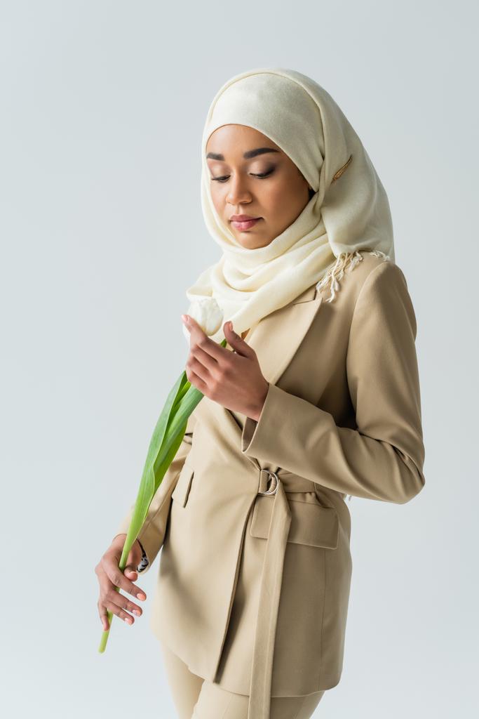 jeune femme musulmane en hijab regardant tulipe isolée sur gris  - Photo, image