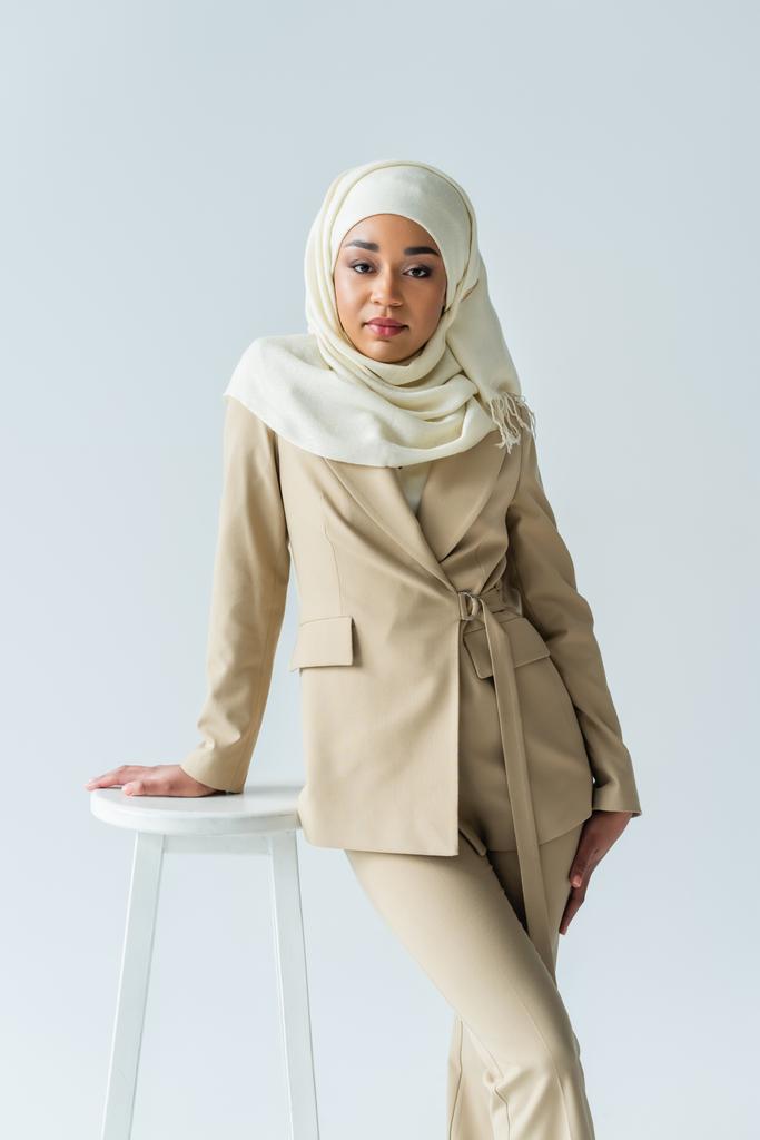 stylowa muzułmanka w hidżabie i garniturze opierająca się na stołku odizolowanym na szarości  - Zdjęcie, obraz