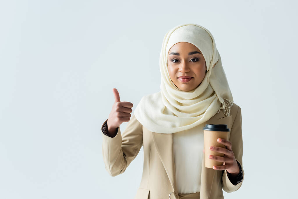 šťastný muslim žena v hidžábu drží papírový pohár a ukazující palec nahoru izolované na šedé - Fotografie, Obrázek
