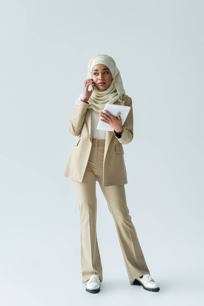 comprimento total da jovem mulher muçulmana no hijab segurando tablet digital e falando no smartphone em cinza - Foto, Imagem