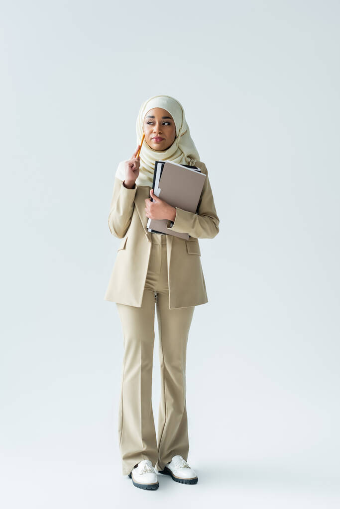 full length of pensive muslim woman in hijab holding pencil and folders on grey - Photo, Image