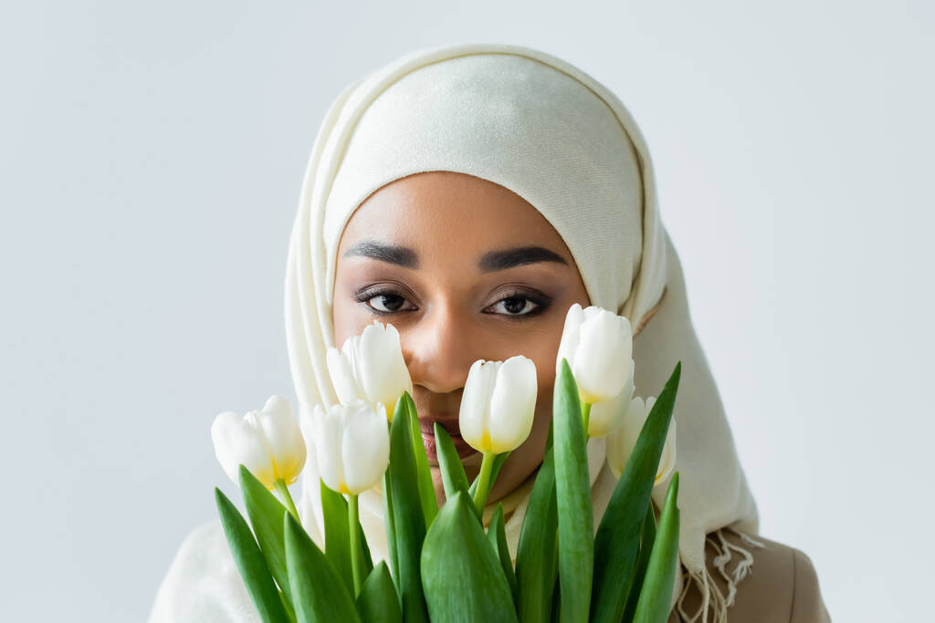 muslim woman in hijab looking at camera through tulips isolated on grey - Photo, Image