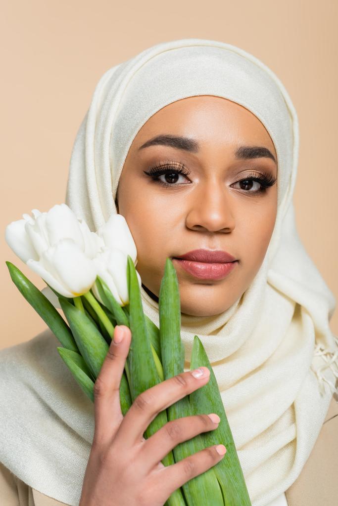 Portrait de jolie femme musulmane en hijab avec maquillage tenant les tulipes isolées sur beige  - Photo, image