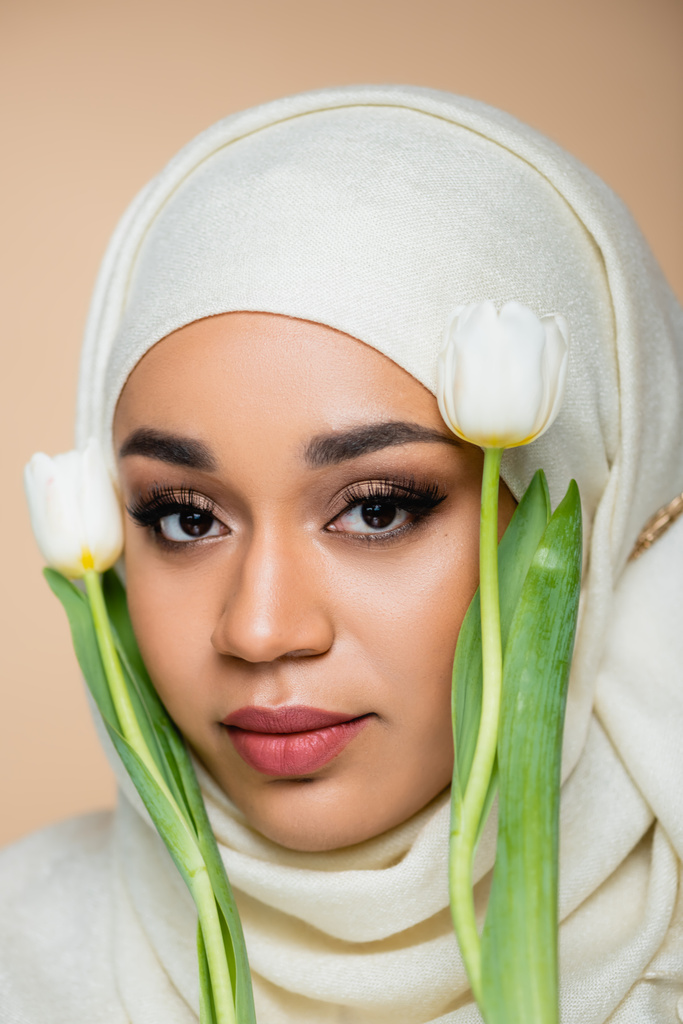 Portret pięknej muzułmanki w hidżabie z makijażem w pobliżu tulipanów wyizolowanych na beżu  - Zdjęcie, obraz