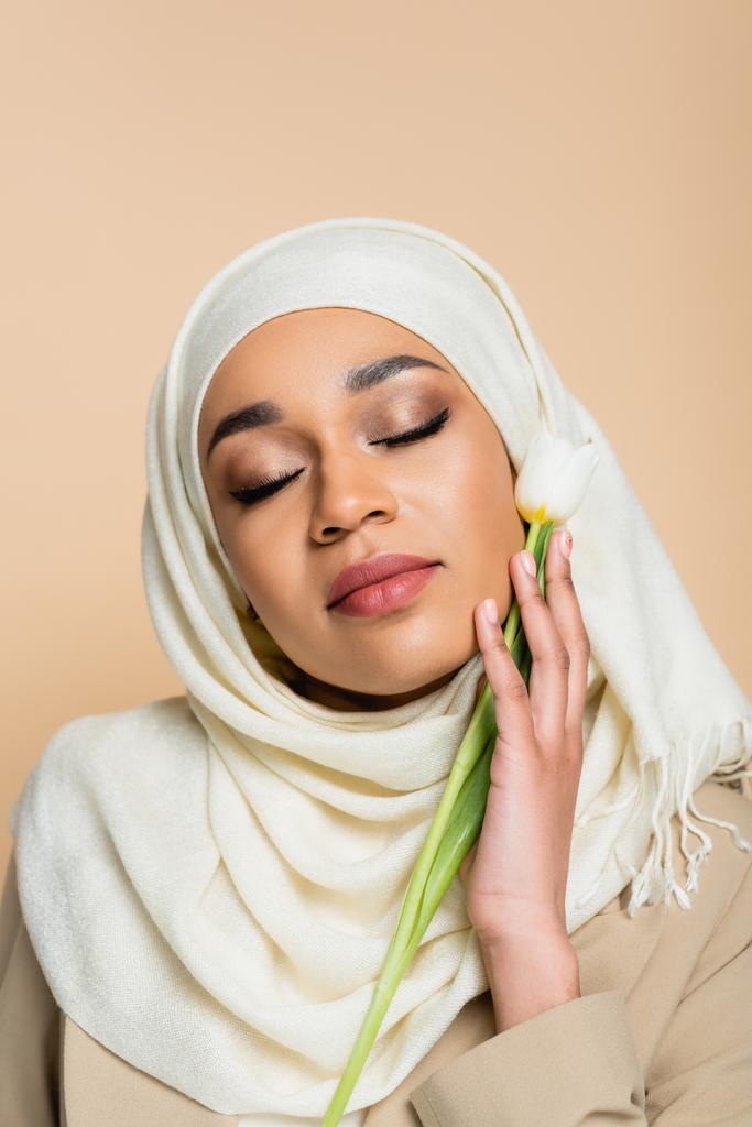 Retrato de una joven musulmana en hijab con los ojos cerrados sosteniendo tulipán aislado en beige  - Foto, imagen