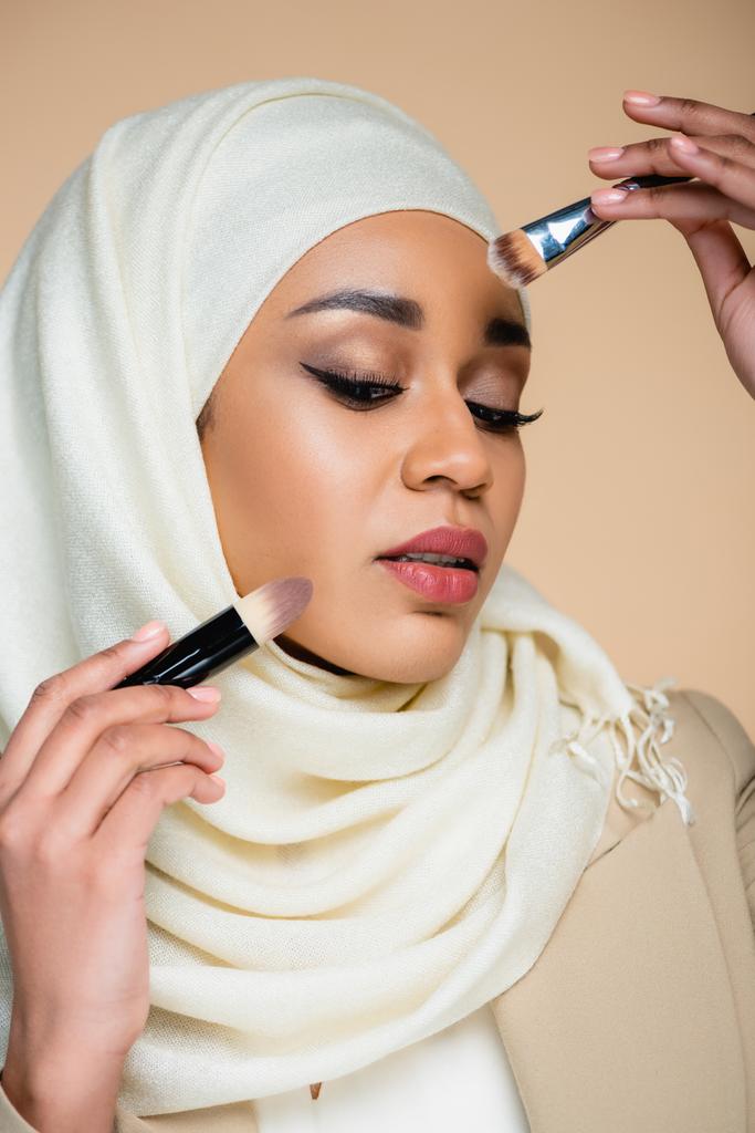 νεαρή μουσουλμάνα γυναίκα σε hijab κρατώντας πινέλα καλλυντικών απομονωμένη σε μπεζ - Φωτογραφία, εικόνα