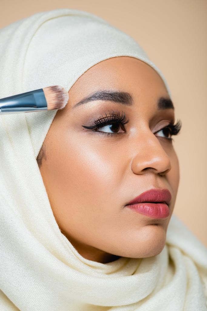retrato de jovem mulher muçulmana em hijab aplicando fundação rosto com escova cosmética isolada em bege - Foto, Imagem