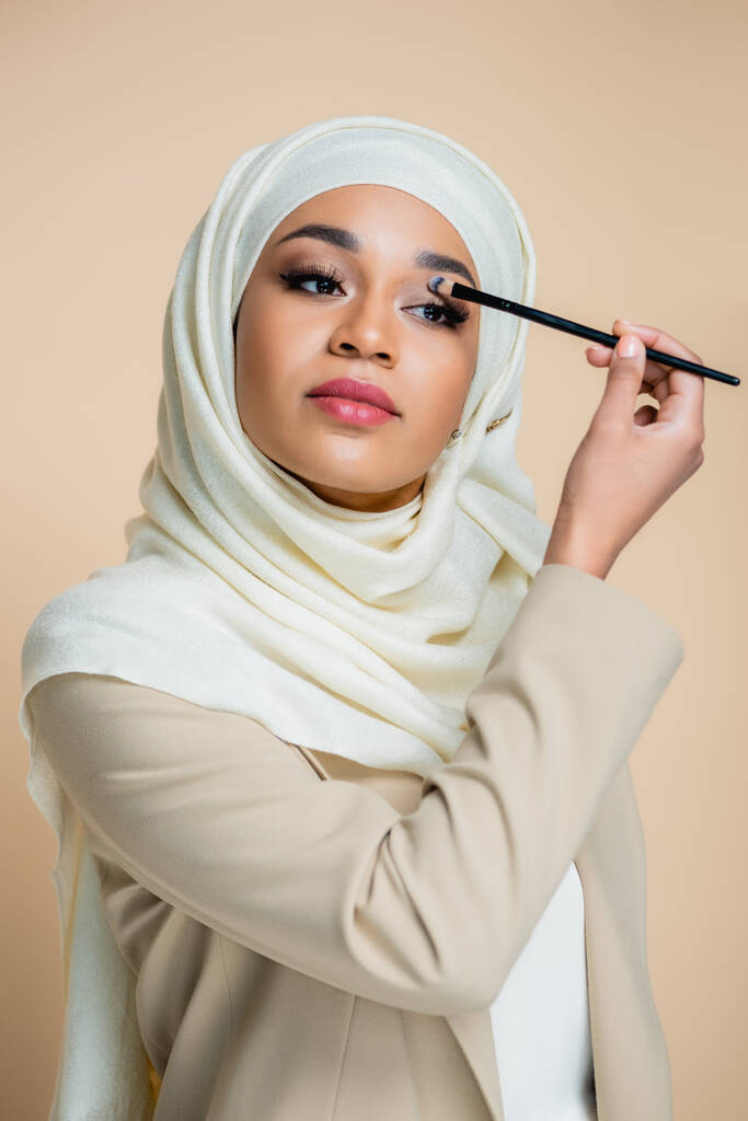young muslim woman in hijab applying eye shadow isolated on beige - Photo, Image