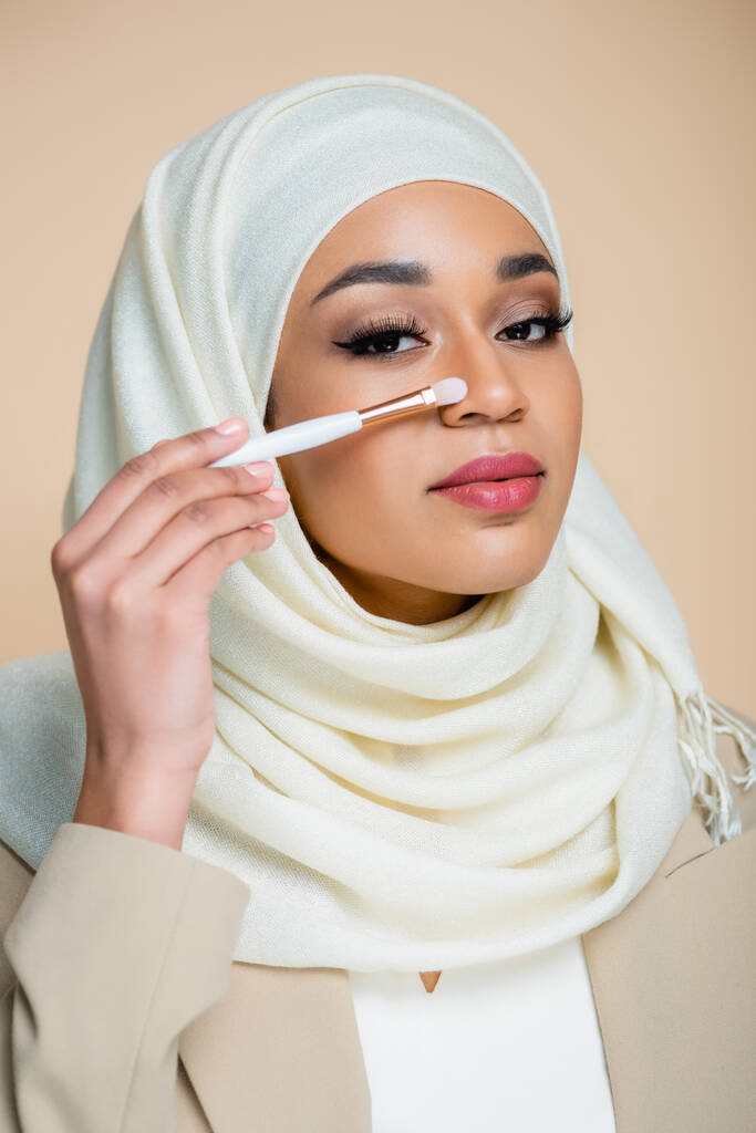 joven mujer musulmana en hijab sosteniendo cepillo cosmético cerca de la cara aislada en beige - Foto, imagen
