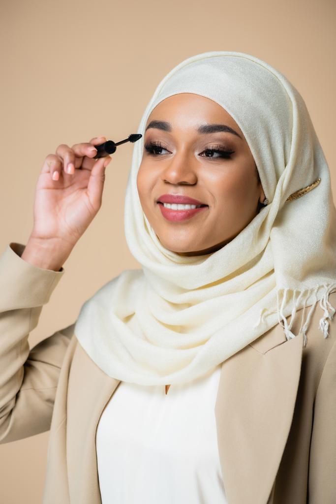 heureuse femme musulmane en hijab appliquant mascara avec pinceau isolé sur beige - Photo, image