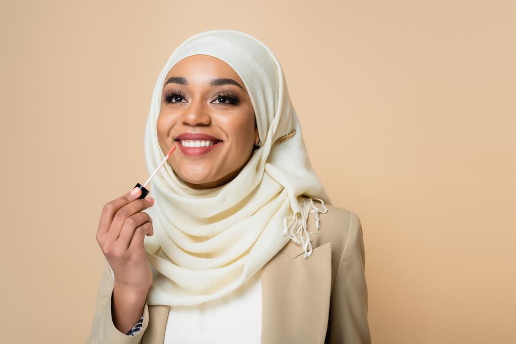 mujer musulmana feliz en hijab aplicando brillo labial aislado en beige - Foto, Imagen