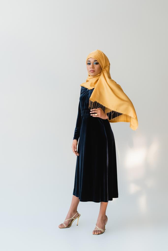 pełna długość marzycielska muzułmanka w hidżabie i aksamitna niebieska sukienka pozująca z ręką na biodrze na białym - Zdjęcie, obraz