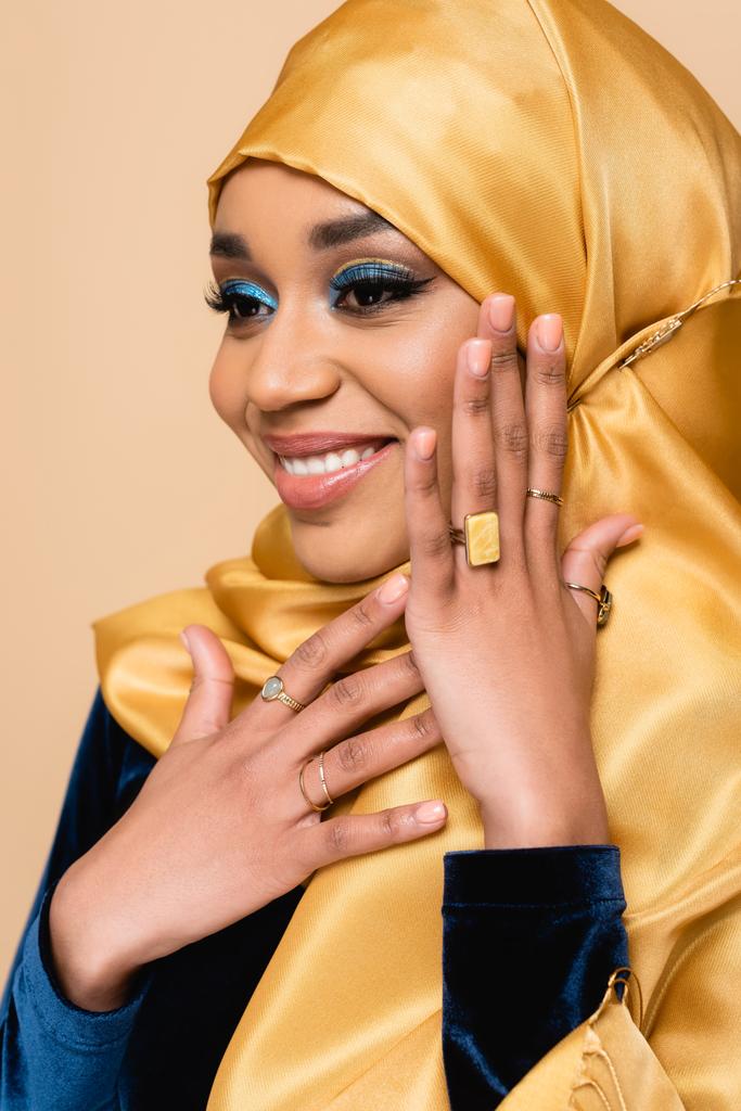 счастливая мусульманка в хиджабе с ярким макияжем, изолированным от бежевого - Фото, изображение