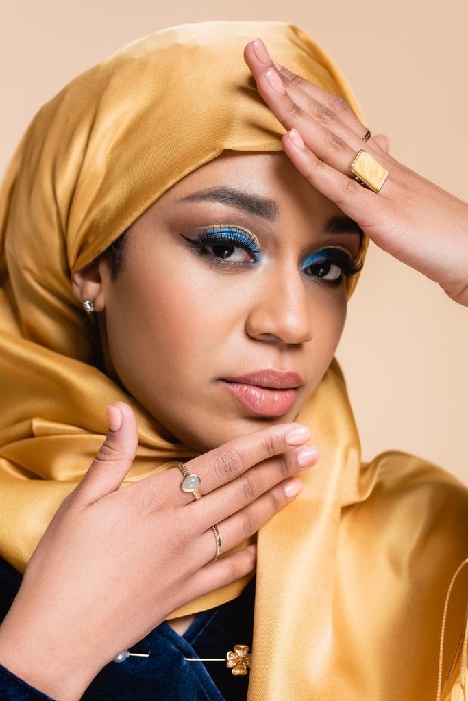 Muslimische Frau in gelbem Hijab mit hellem Make-up und goldenen Ringen blickt vereinzelt in die Kamera auf beige - Foto, Bild