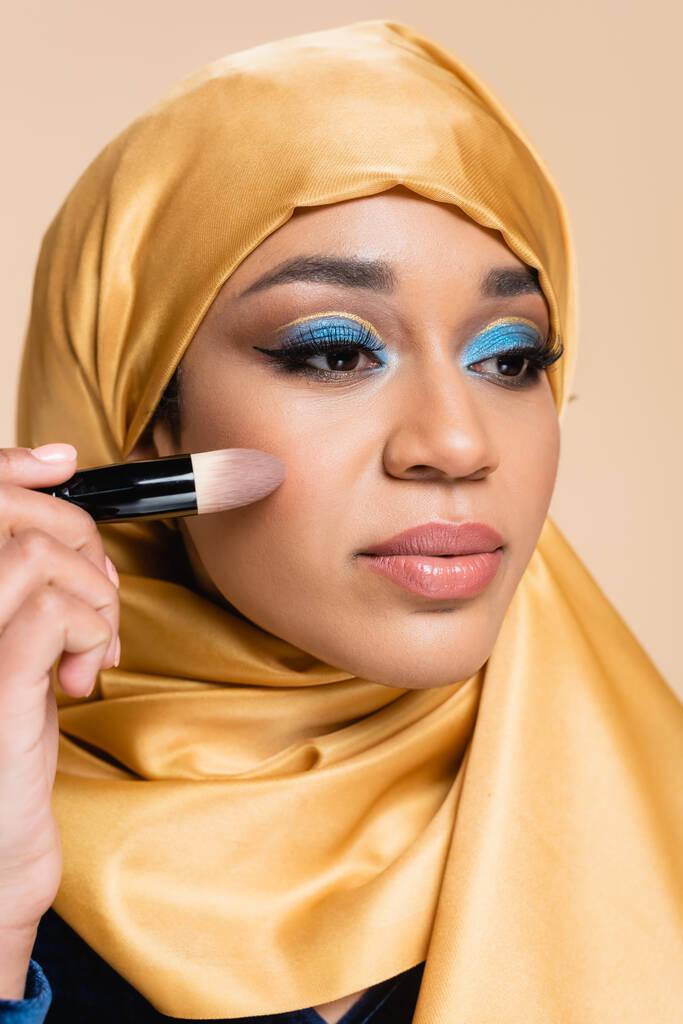 jonge moslim vrouw in gele hijab aanbrengen make-up foundation met cosmetische borstel geïsoleerd op beige - Foto, afbeelding