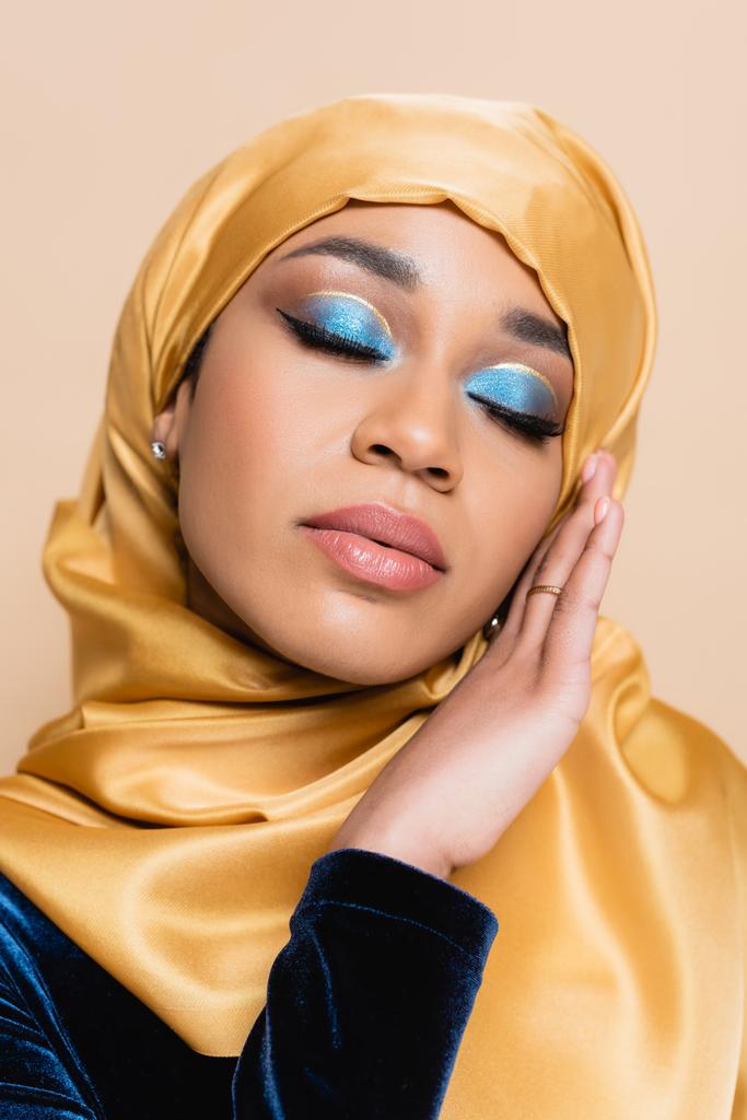 muslim γυναίκα σε μαντίλα με λαμπερό μπλε μακιγιάζ ματιών και κλειστά μάτια απομονώνονται σε μπεζ - Φωτογραφία, εικόνα