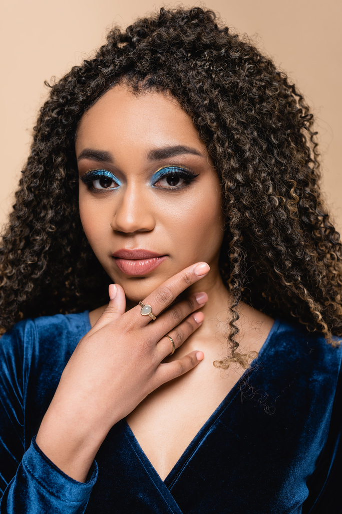 curly african american woman with makeup touching chin isolated on beige - Photo, Image
