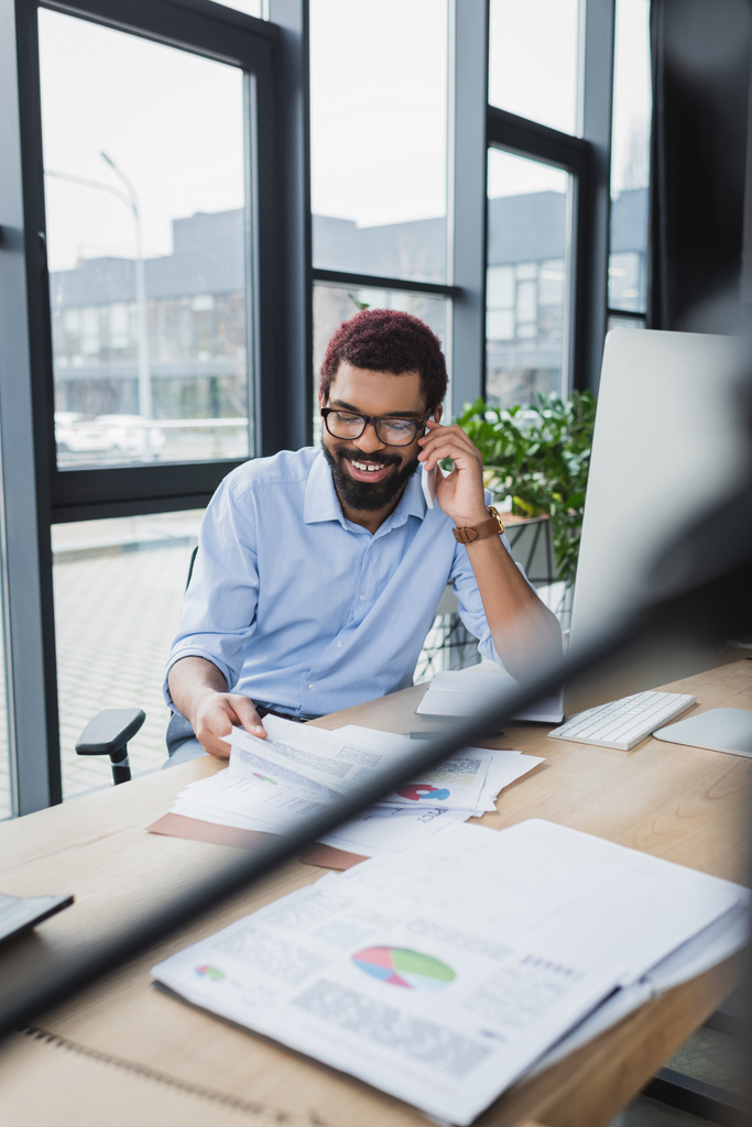 Θετικός Αφροαμερικάνος επιχειρηματίας που μιλάει σε smartphone και κρατάει χαρτί με διαγράμματα στο γραφείο  - Φωτογραφία, εικόνα