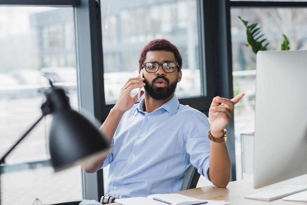 Uomo d'affari afroamericano che parla su smartphone e punta il dito contro il computer in ufficio  - Foto, immagini