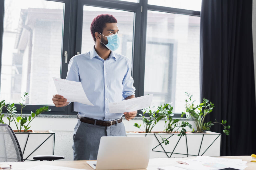 Αφροαμερικάνος επιχειρηματίας με ιατρική μάσκα που κρατάει χαρτιά κοντά σε λάπτοπ στο γραφείο  - Φωτογραφία, εικόνα