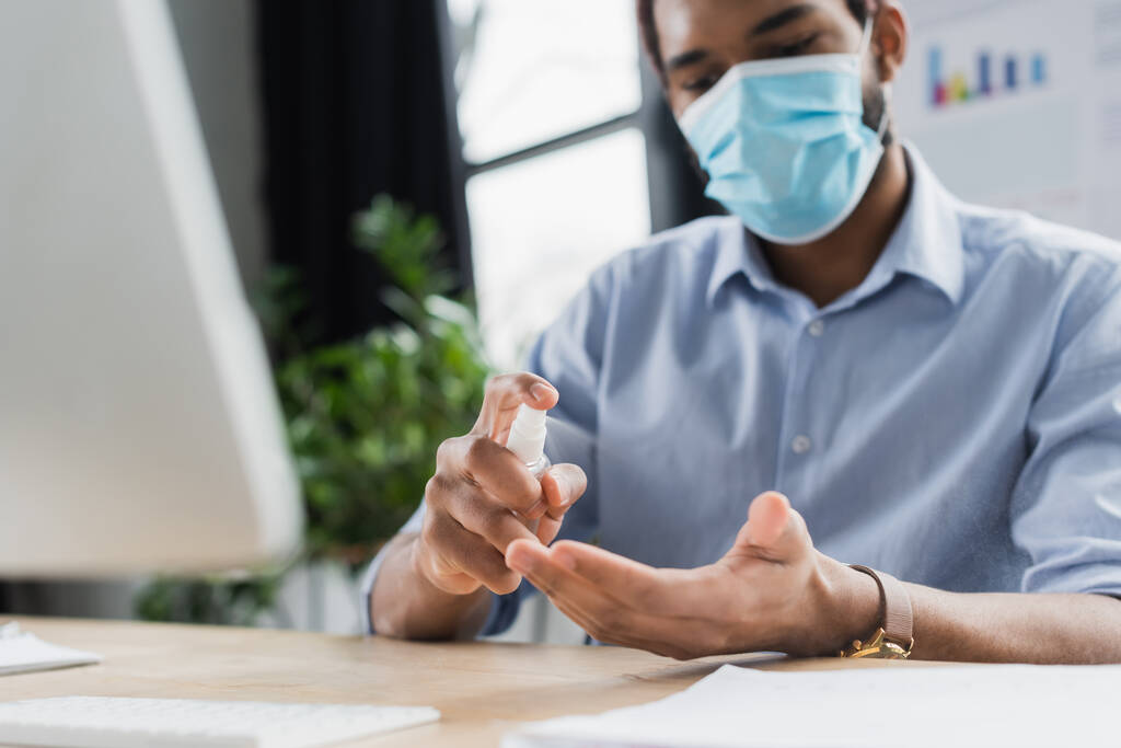 Bulanık Afro-Amerikan iş adamı tıbbi maskeli. Ofisteki bilgisayarın yanında el dezenfektanı sıkıyor.  - Fotoğraf, Görsel