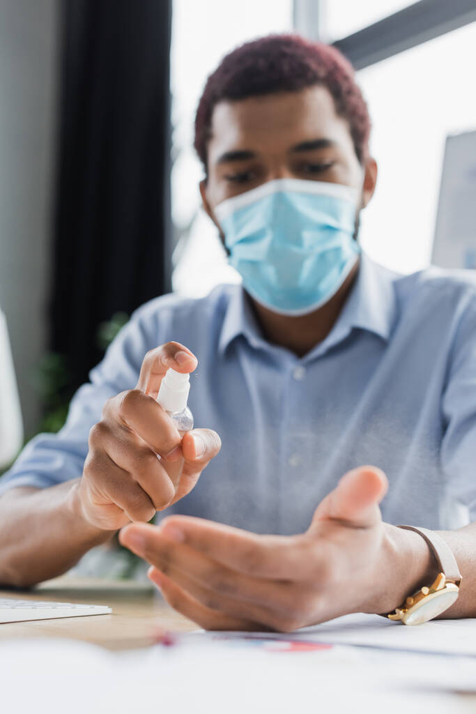 Gerente afroamericano borroso en mascarilla médica rociando desinfectante de manos en la oficina  - Foto, imagen