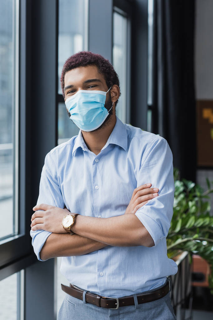 Homme d'affaires afro-américain en masque médical debout avec les bras croisés au bureau  - Photo, image