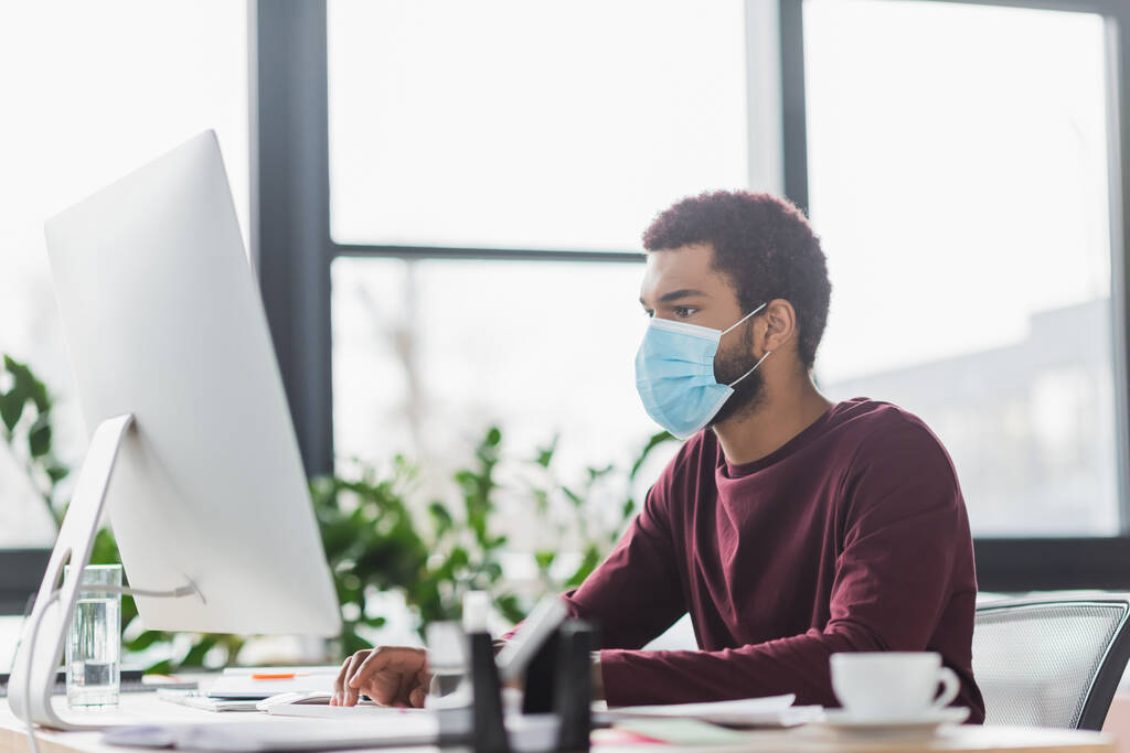 African american businessman in medical mask using computer near coffee and water in office  - Photo, Image