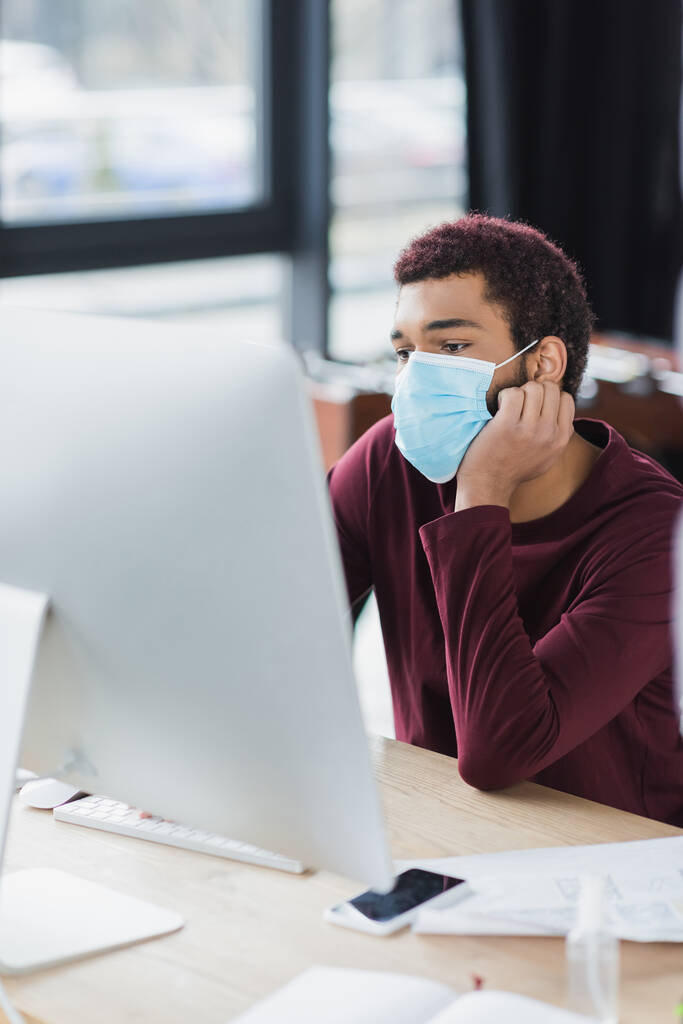 Hombre de negocios afroamericano en máscara médica mirando el monitor de la computadora cerca de teléfono inteligente y desinfectante de manos - Foto, Imagen