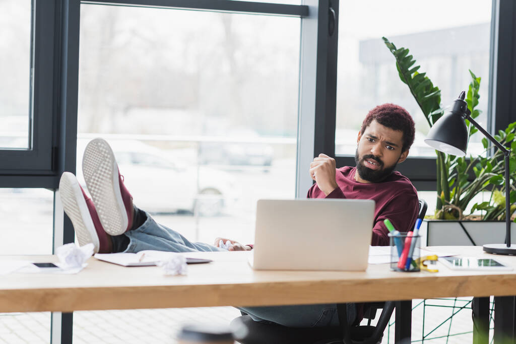 Boze Afro-Amerikaanse zakenman op zoek naar laptop in de buurt van verkreukeld papier in het kantoor  - Foto, afbeelding