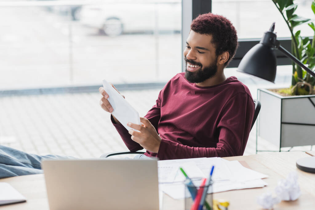 Sonriente hombre de negocios afroamericano utilizando tableta digital cerca de la computadora portátil y papel arrugado en la oficina  - Foto, imagen