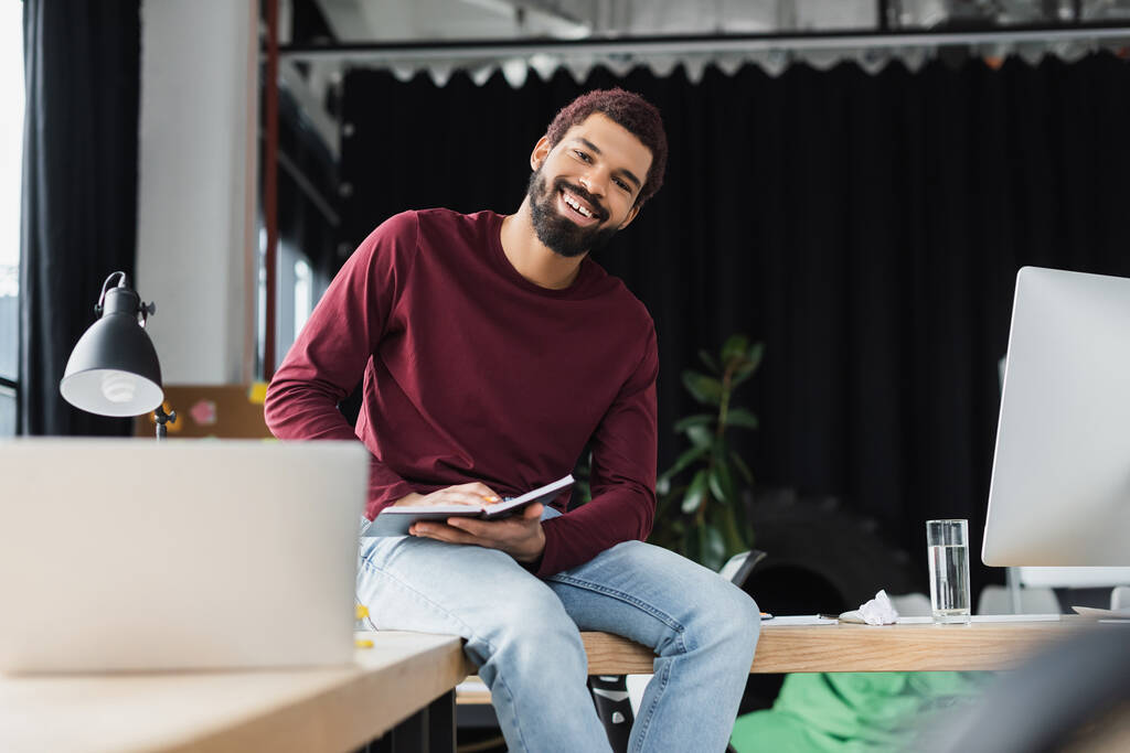 Empresário afro-americano sorrindo para a câmera enquanto segurava notebook perto do laptop no escritório  - Foto, Imagem